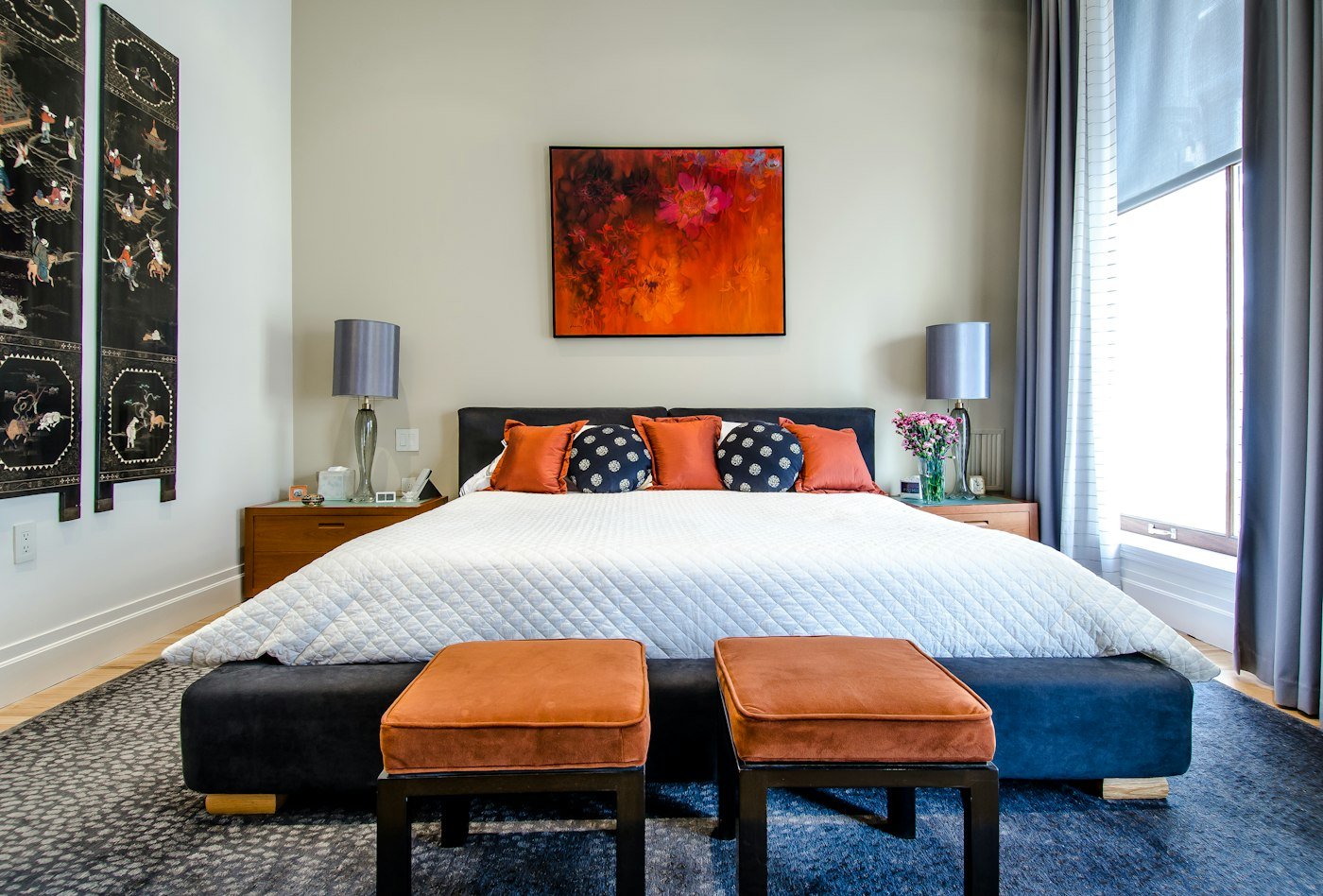 A white quilted bed, orange cushions, a bright floral painting, and two rust stools